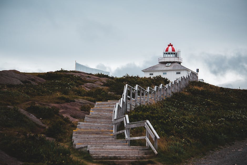 A winding wooden staircase with weathered, greyish planks and simple railings ascends the grassy hillside towards a white lighthouse with a red dome and lantern room, set against a cloudy sky. The hil